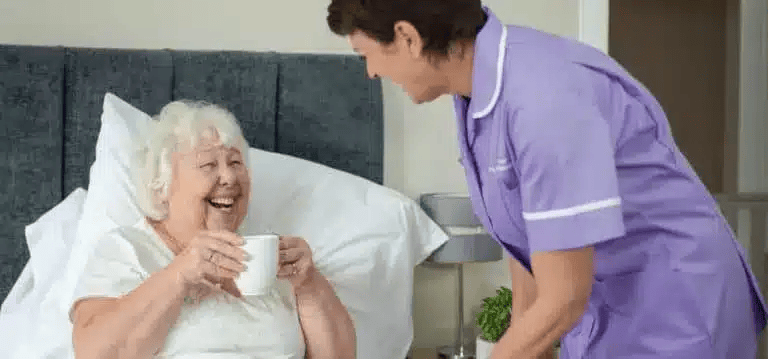 Elderly-lady-in-bed-with-hot-drink-female-carer-purple-uniform_2x1-1-768x359-1.png